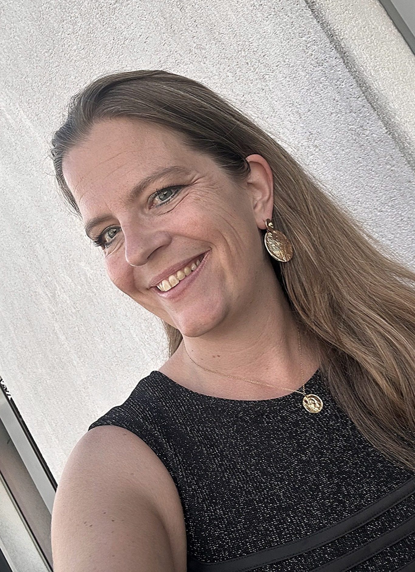 Gabriela De Forrest- Autorin Eine lächelnde Frau mit langen, dunkelblonden Haaren und Ohrringen. Sie trägt ein schwarzes Oberteil. Das ist die Autorin.